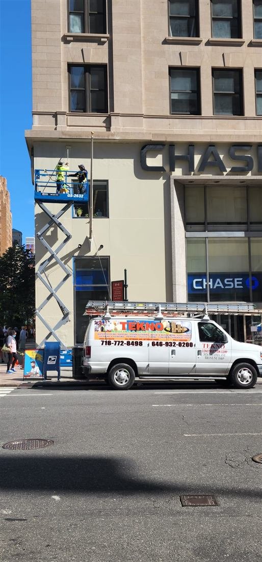 Wide sunny street view of the same Chase branch facade job: crew on a raised scissor lift with the fully branded Tekno Construction van parked below - strong commercial-credibility shot.