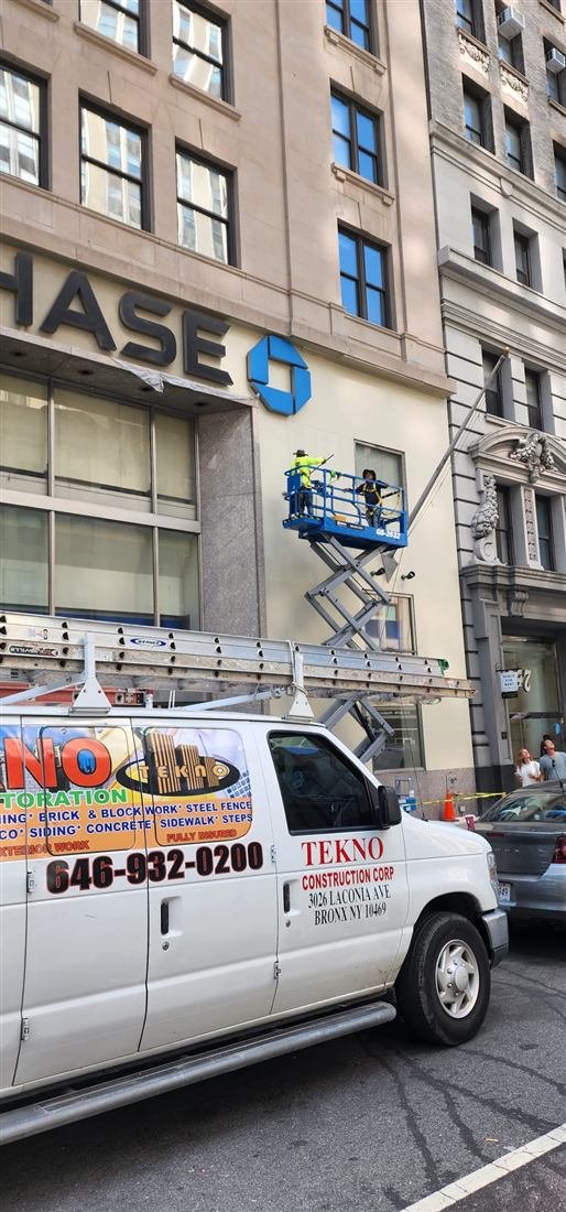 Two workers on a Genie scissor lift restoring/painting the facade of a Chase bank branch in Manhattan with the branded Tekno van and ladder rack in the foreground.