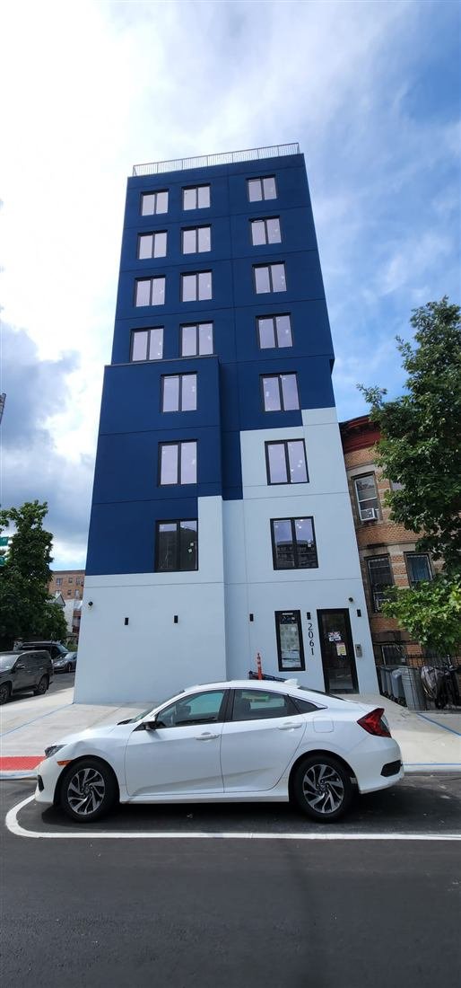 Street-level shot of a completed narrow new-construction apartment building (2061) with striking two-tone navy and white facade in a residential NYC neighborhood; parked car dominates the foreground.