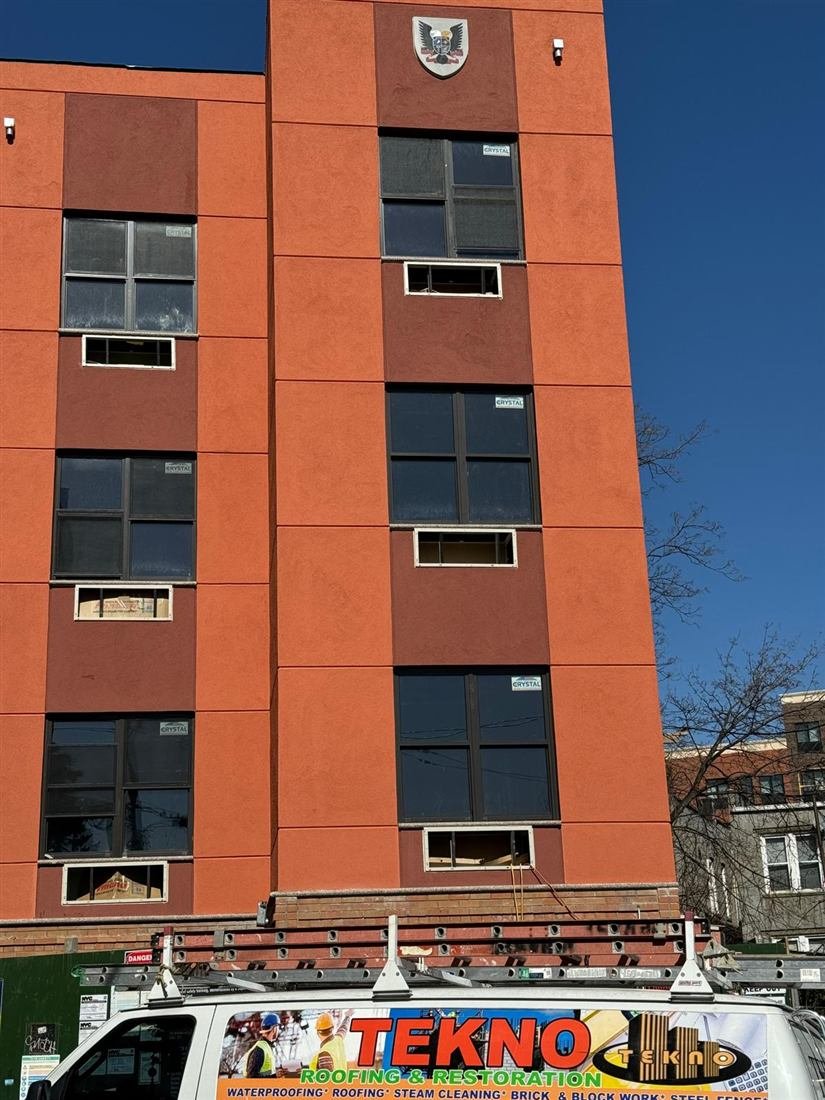 Multi-story orange/red stucco commercial-residential building facade with newly installed windows still carrying manufacturer stickers, Tekno Roofing & Restoration branded van parked in the foreground.