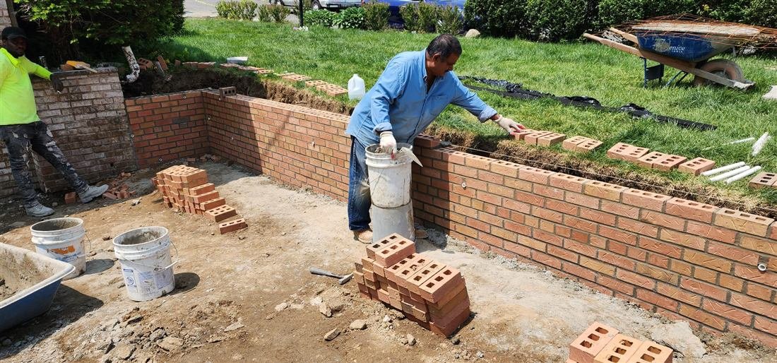 Two masons actively building a brick retaining/foundation wall at a residential front yard, with brick stacks, mortar buckets and a wheelbarrow — one worker's face is clearly identifiable, so get consent before publishing.