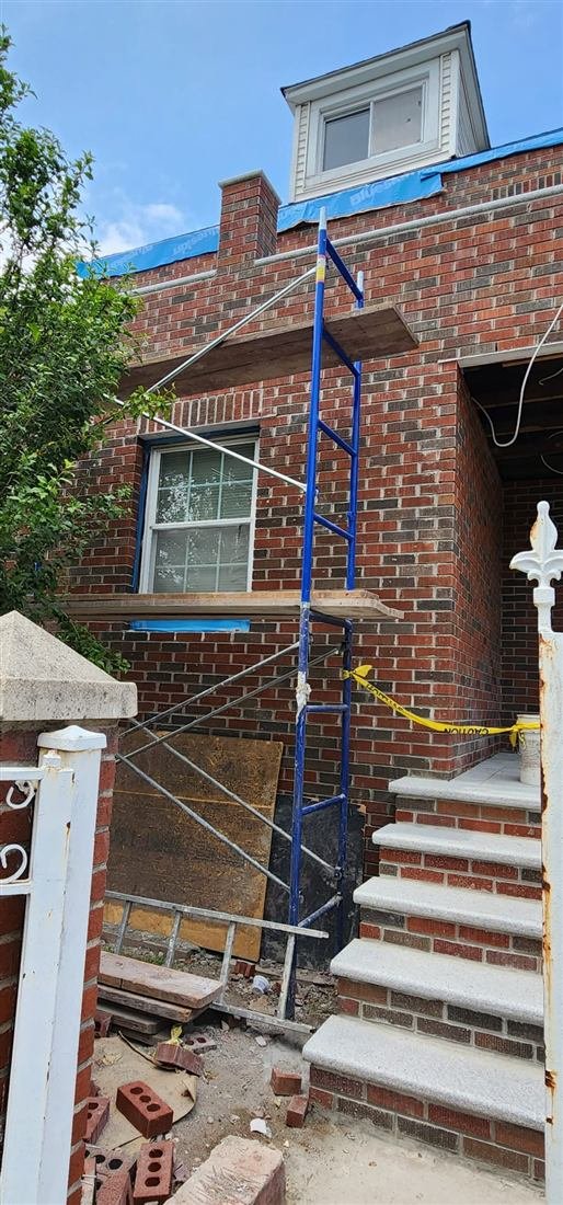 Frame scaffolding erected against a brick residential house during masonry/brick repair, with new granite steps, caution tape, and loose bricks staged at grade.