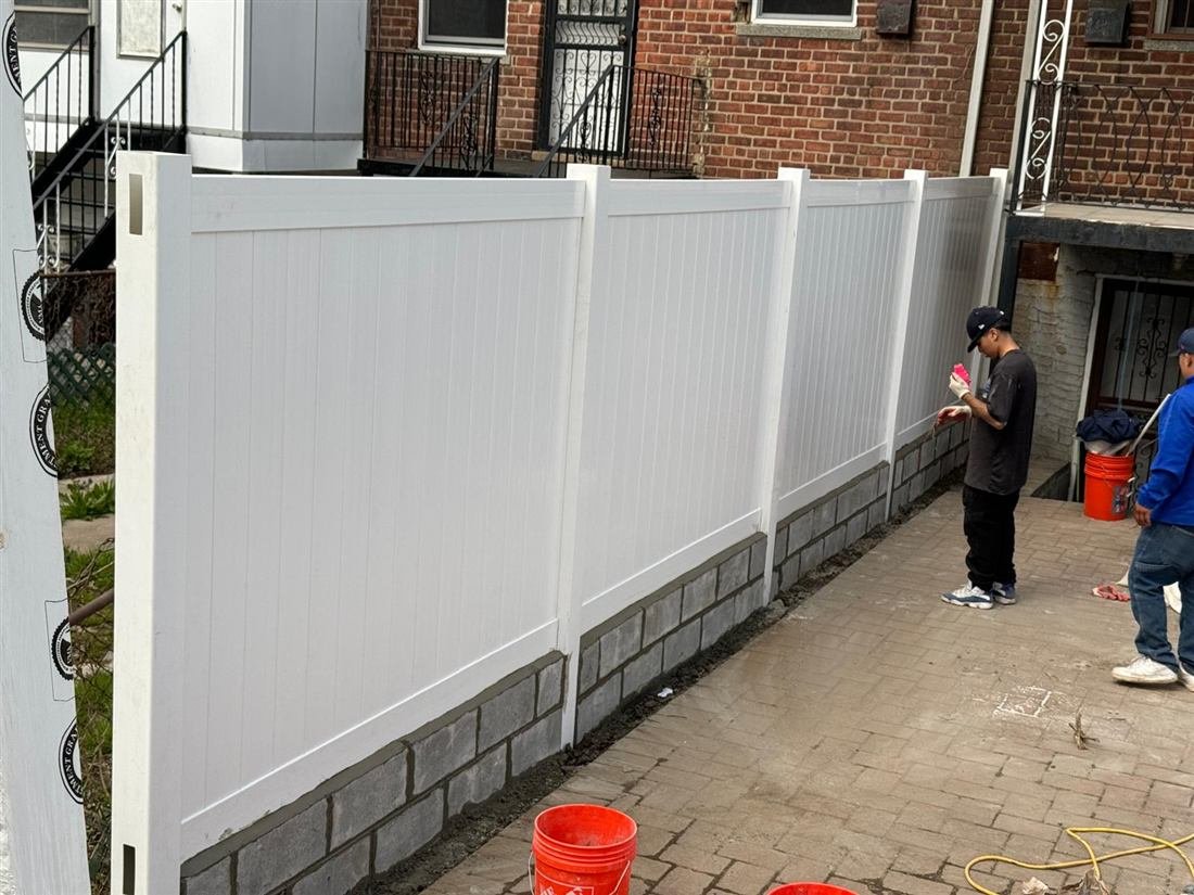 New white vinyl privacy fence being installed on a fresh cinder-block knee wall in a residential backyard with brick-paved patio; two workers are finishing the mortar joints.
