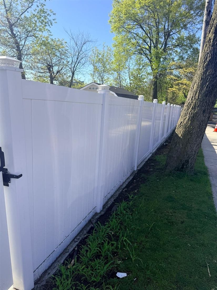 Long run of newly installed white vinyl privacy fence with decorative post caps along a residential sidewalk and grass strip, bright sunny conditions.