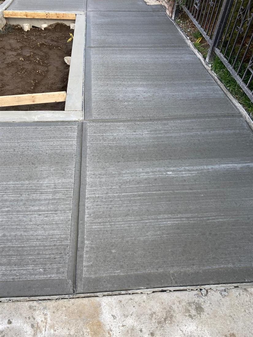 Freshly finished broom-textured concrete sidewalk flags with a formed tree pit and wrought-iron fence along a residential street.
