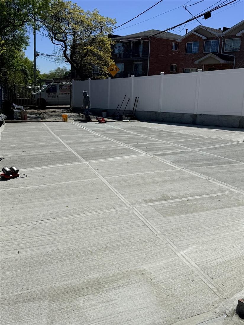 Large freshly finished broom-scored concrete driveway/parking pad at a residential property with new white PVC fence, Tekno branded van and one worker in the background.
