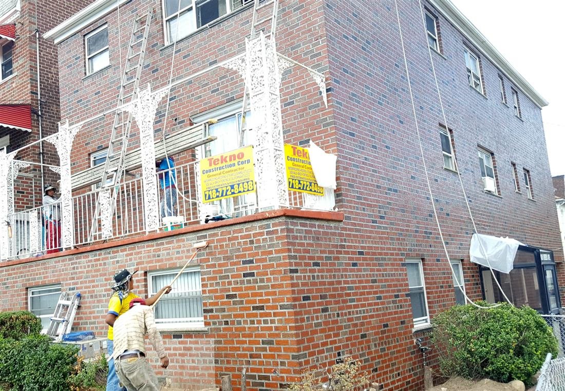 Crew waterproofing/coating the brick facade of a residential corner building with ladders and roller poles, two Tekno Construction Corp yard signs prominently displayed.