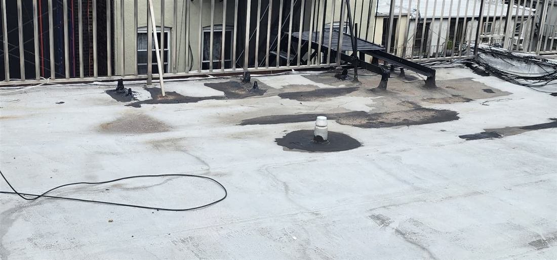 Flat residential roof mid-repair with black mastic patches around pipe penetrations and equipment supports over a worn silver coating — documentary in-progress shot, cluttered with cables.